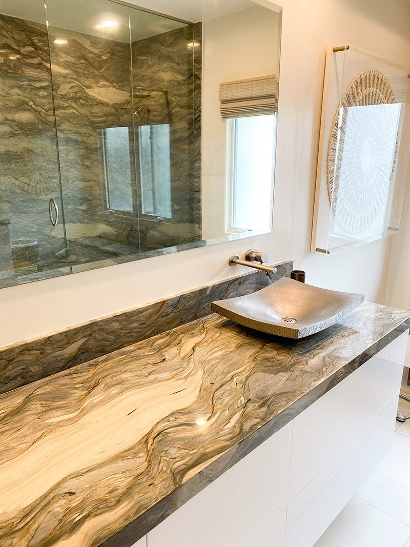 Prefabricated stone vanity countertop and backsplash with flowing gold, cream and gray veining, a brushed nickel vessel sink and wall-mount faucet on a white floating vanity